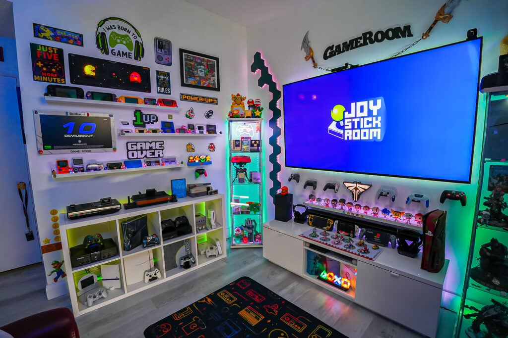 Gaming showcase featuring PlayStation, Xbox, Atari, and Nintendo consoles, display case with collectible figures, controller collection, modular LED lights, and video game posters
